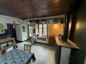 Eine Küche mit Holzbalkendecke, einem Esstisch mit gemustertem Tischläufer, einem Stuhl und einem Fenster mit Blick ins Grüne.