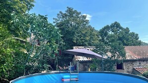 En swimmingpool med ett blått överdrag, omgiven av träd och en byggnad i sten med ett tak täckt av tegelpannor.