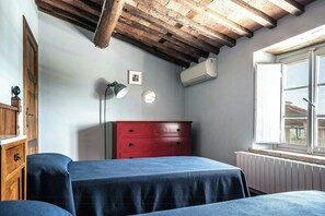 Ein Zimmer mit Holzbalkendecke, einem roten Schminktisch, einem Bett mit blauer Tagesdecke, einer Wand-Klimaanlage und einem Fenster mit Blick.