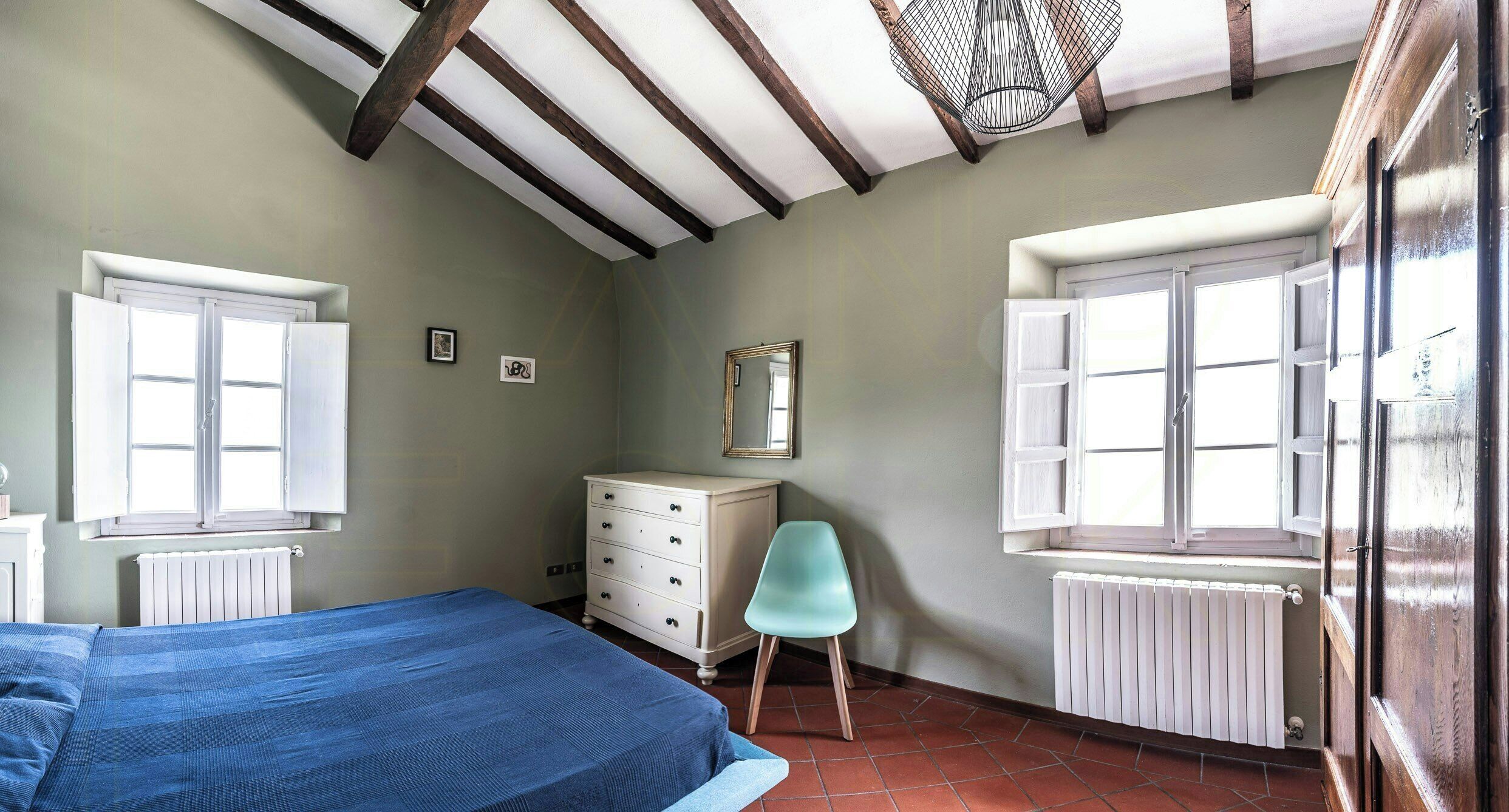 Une chambre à coucher comprenant un lit, une commode, une chaise et deux fenêtres équipées de volets.