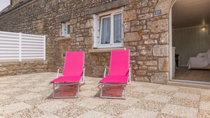 A stone building with a number '20' sign, a window with lace curtains, and two pink lounge chairs outside.