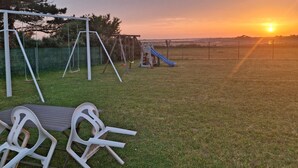 A grassy outdoor area with a swing set and a slide, a picnic table, and a view of the sunset.