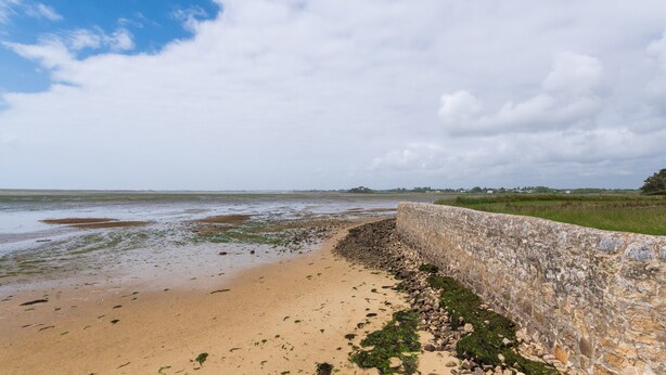 A coastal landscape with a stone wall, sandy beach, and distant greenery.