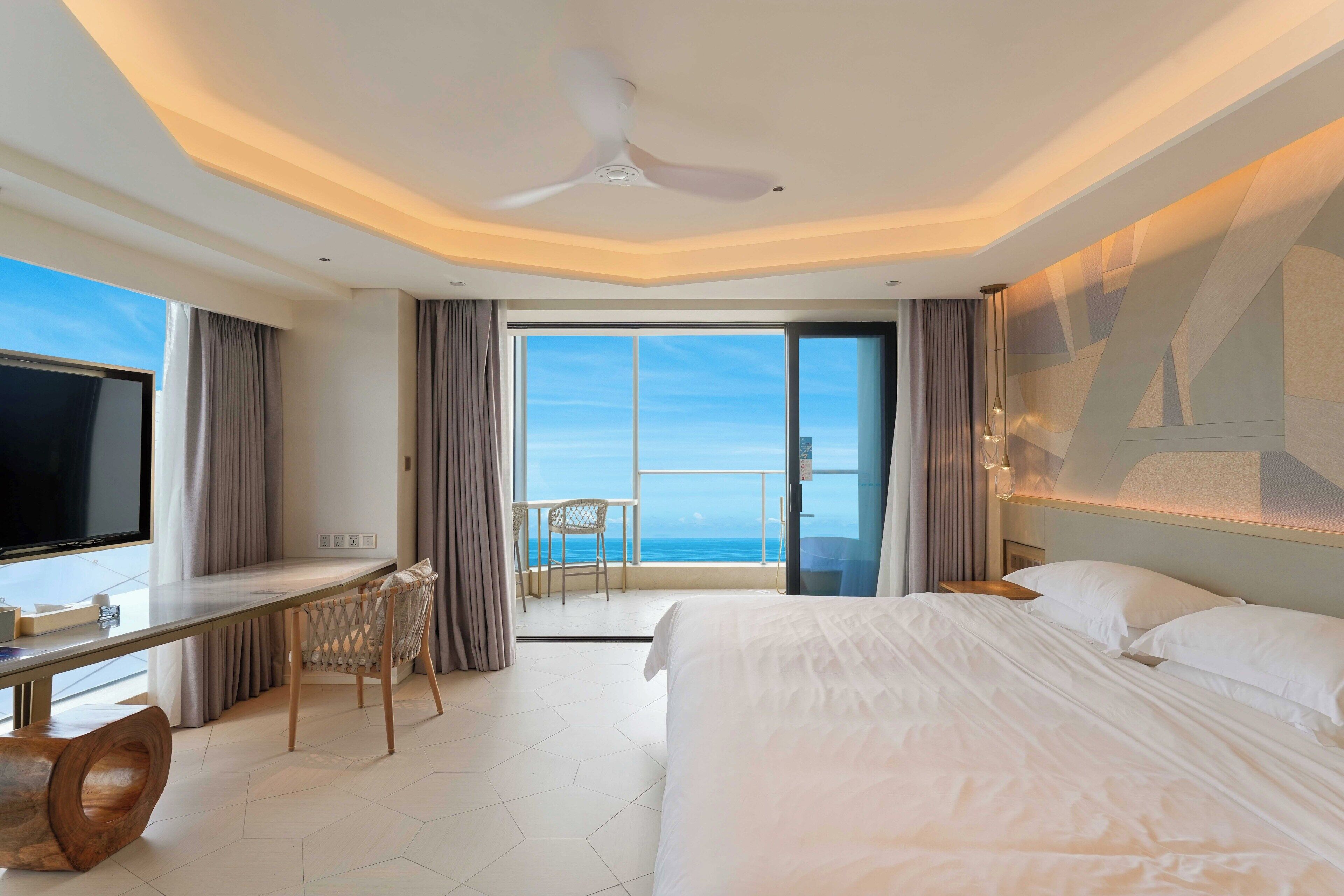 Una habitación de hotel con cama, escritorio, silla, televisor y balcón con vista al océano.