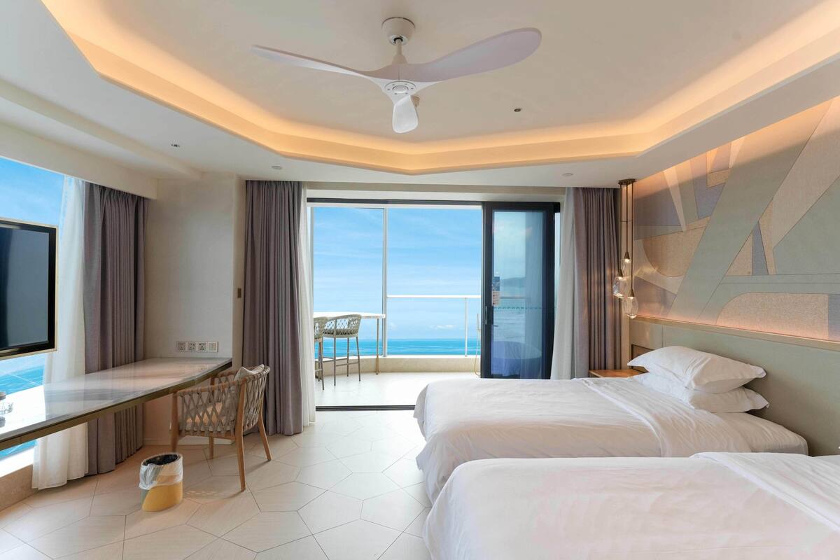 A hotel room with a bed, a desk, a chair, a TV, and a balcony with a view of the ocean.