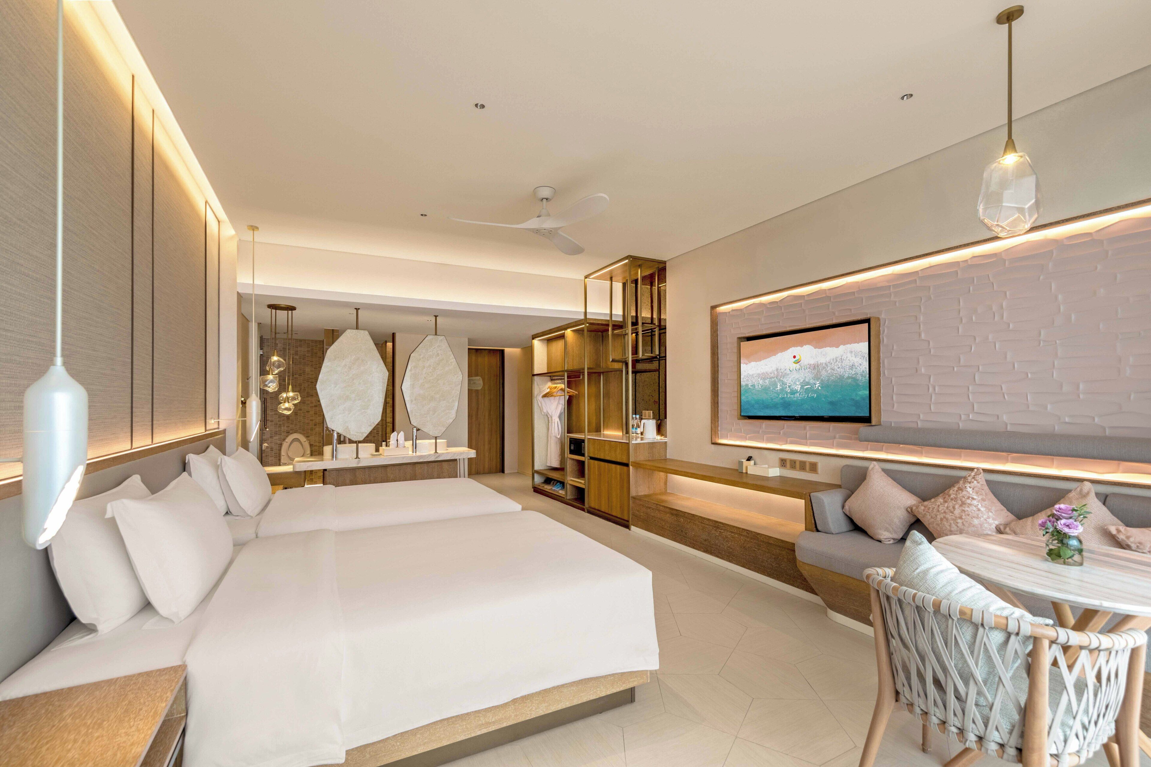 A modern hotel room with a large bed, a dining area, a TV mounted on the wall, and a ceiling fan.