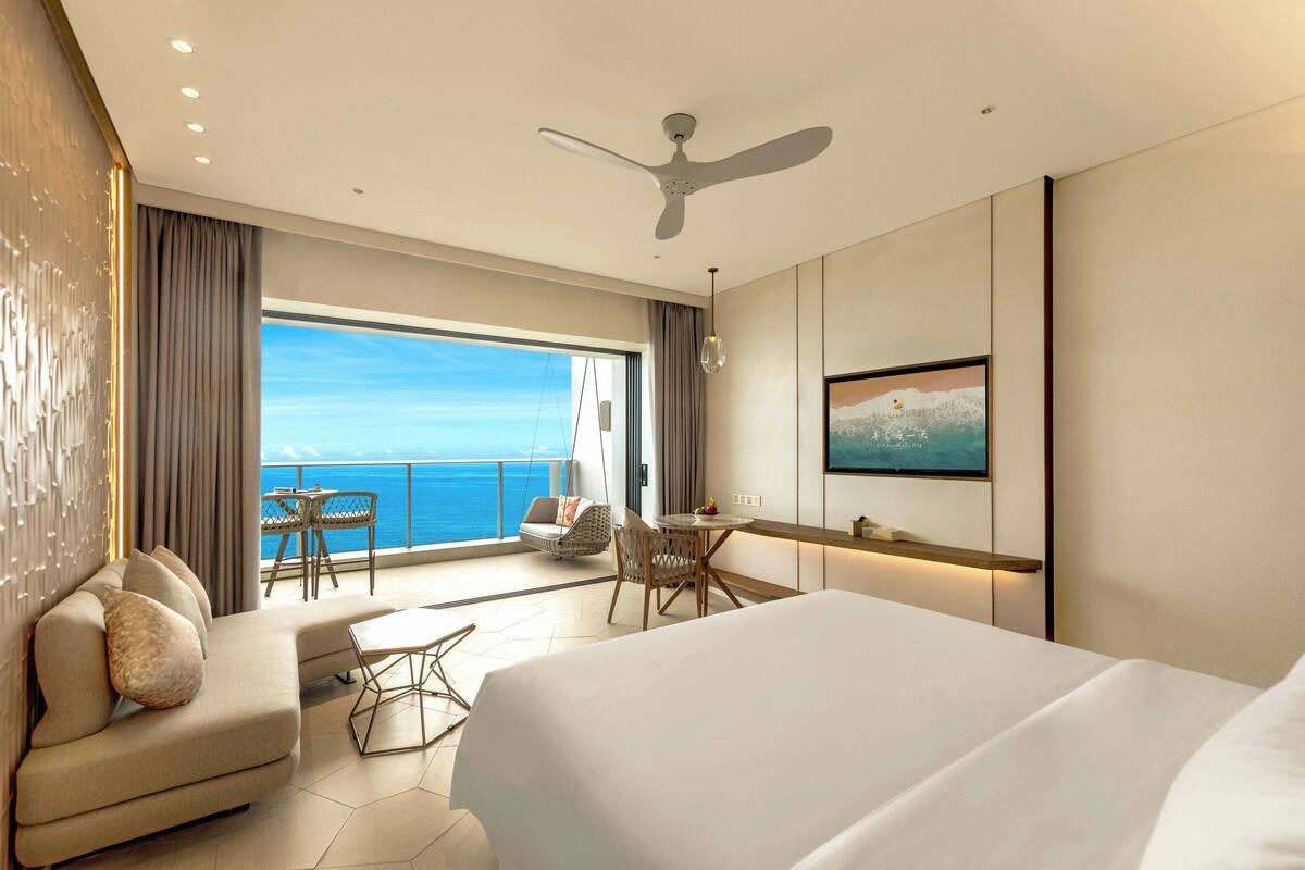A hotel room with a large bed, a sofa, a dining table, a ceiling fan, and a view of the ocean.