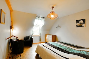 Une chambre à coucher comprenant un lit, une commode en bois, une chaise, un luminaire suspendu et une fenêtre avec des rideaux.