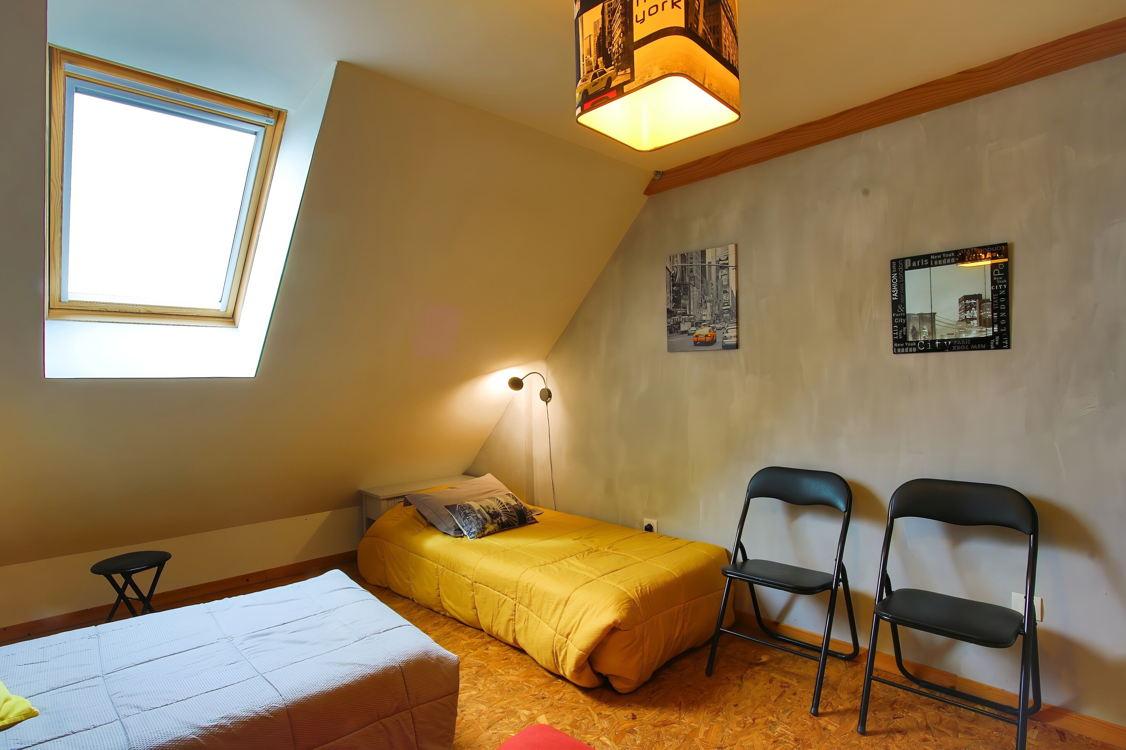 Um apartamento pequeno e de um único quarto, com teto inclinado, claraboia, cama, duas cadeiras e duas imagens penduradas na parede.