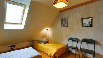 Um apartamento pequeno e de um único quarto, com teto inclinado, claraboia, cama, duas cadeiras e duas imagens penduradas na parede.