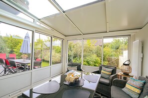 Ein Wintergarten mit großen Fenstern, einem Tisch, Stühlen und einem Korbstuhl.