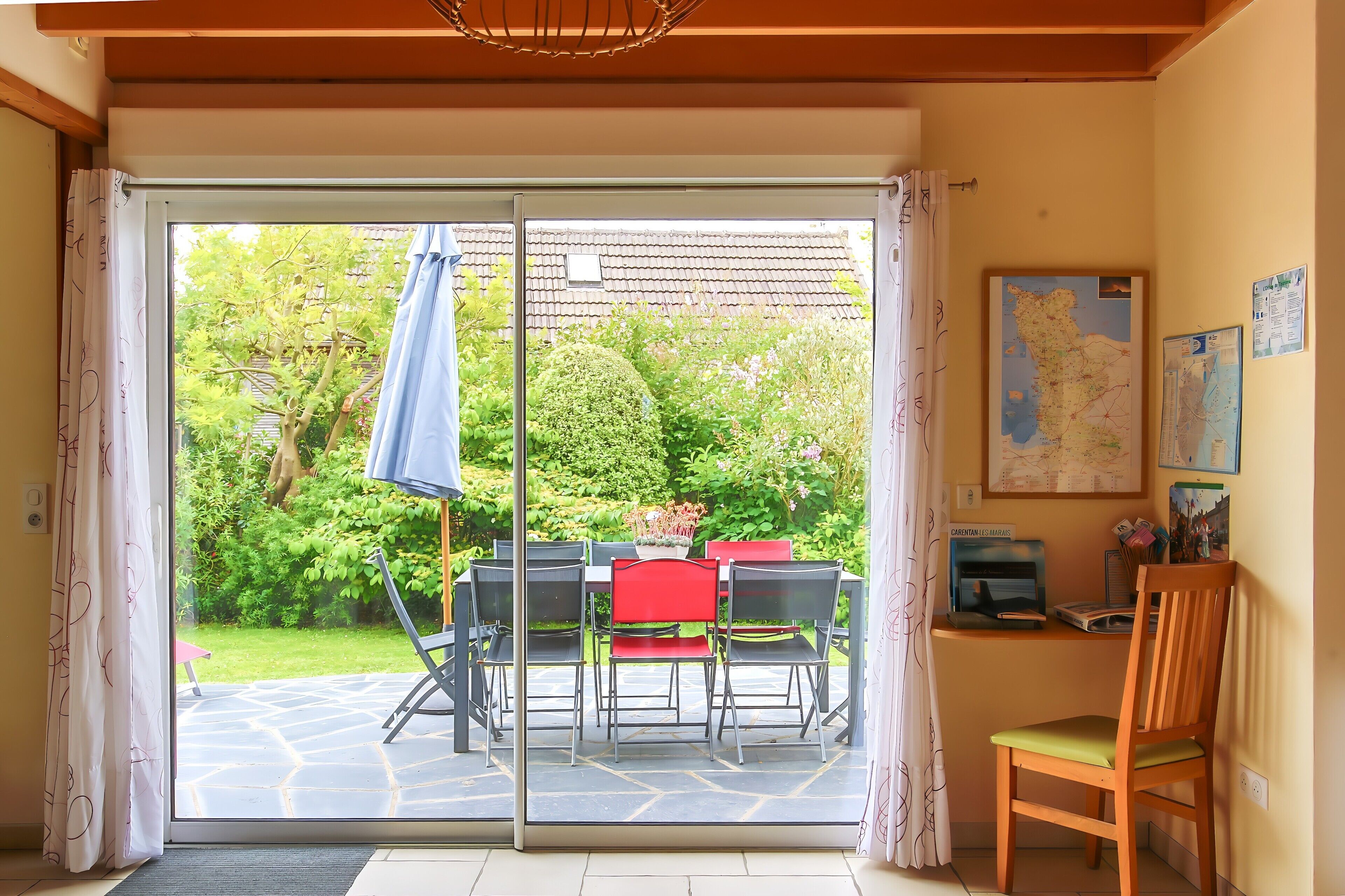 A room with a sliding glass door leading to a garden, a table with chairs, a wooden chair, and a small desk with a map and books.
