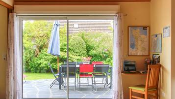 A room with a sliding glass door leading to a garden, a table with chairs, a wooden chair, and a small desk with a map and books.
