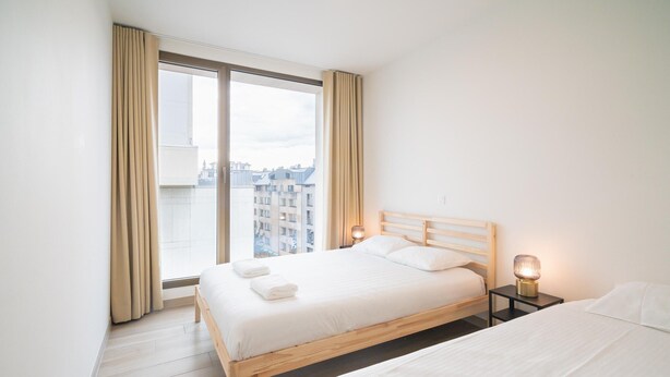 A hotel room with a large window, a wooden bed, a bedside table with a lamp, and a second bed. - Opera Square Family Apartment for 10 Guests (Antwerp)