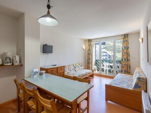 Ein Zimmer mit Esstisch, Stühlen, einer Couch, einem Fernseher und einem Balkon mit Vorhängen.
