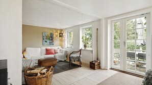 A spacious living area with a white sofa, a wooden chair, a wicker basket, and a view through large glass doors to an outdoor patio. - Nature-close home near beach at Limfjorden (Thyholm)