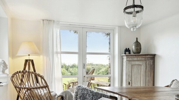 A cozy room with a wooden dining table, wicker chairs, a rocking chair, and a view through large windows. - Nature-close home near beach at Limfjorden (Thyholm)