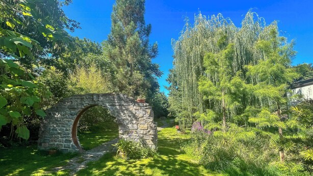 Un arc en pierre se dresse dans un jardin luxuriant, où poussent différentes espèces d’arbres et de plantes.