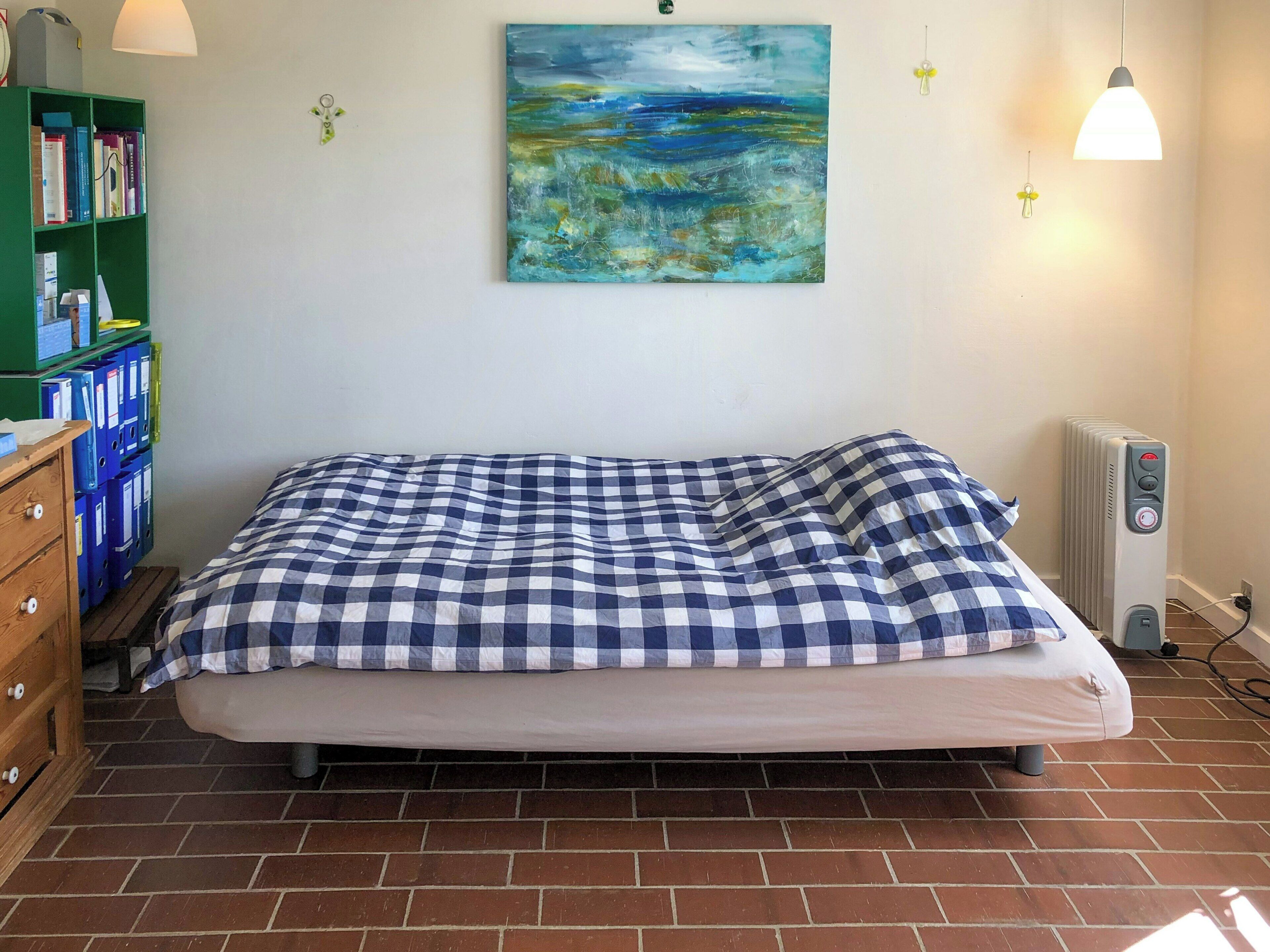 Un lit recouvert d’une couverture à carreaux, un radiateur, une bibliothèque et un tableau accroché au mur.