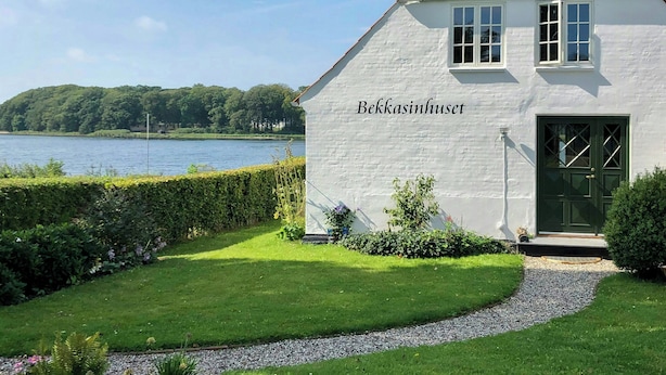 Ein weißes Gebäude mit einer grünen Tür und Fenstern, mit dem Schriftzug 'Bekkasinhuset', mit Blick auf einen See und Hügel im Hintergrund.