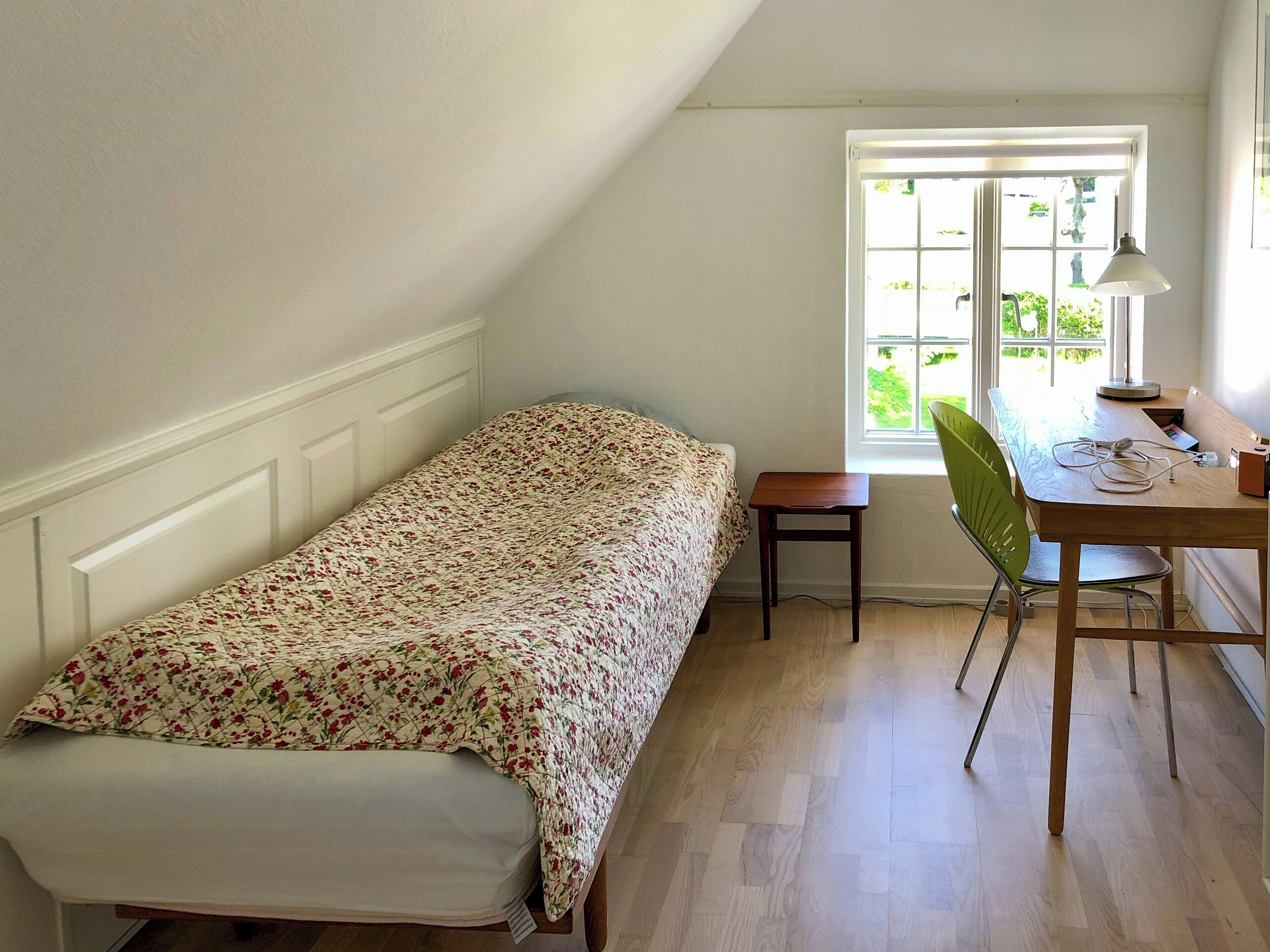Una habitación pequeña con cama individual, un escritorio y una silla de madera, una ventana con vista a zonas verdes y un radiador en la pared.