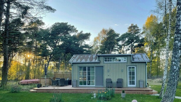 A small, single-story building with a flat roof, large windows, and a patio area with outdoor furniture.