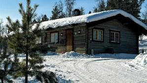 A log cabin with a snow-covered roof, surrounded by snow and pine trees.