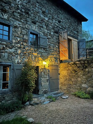 A stone building with a wooden door and shuttered windows, surrounded by gravel and greenery. - Architect's house in an Auvergne hamlet (Saint-Martin-des-Olmes)