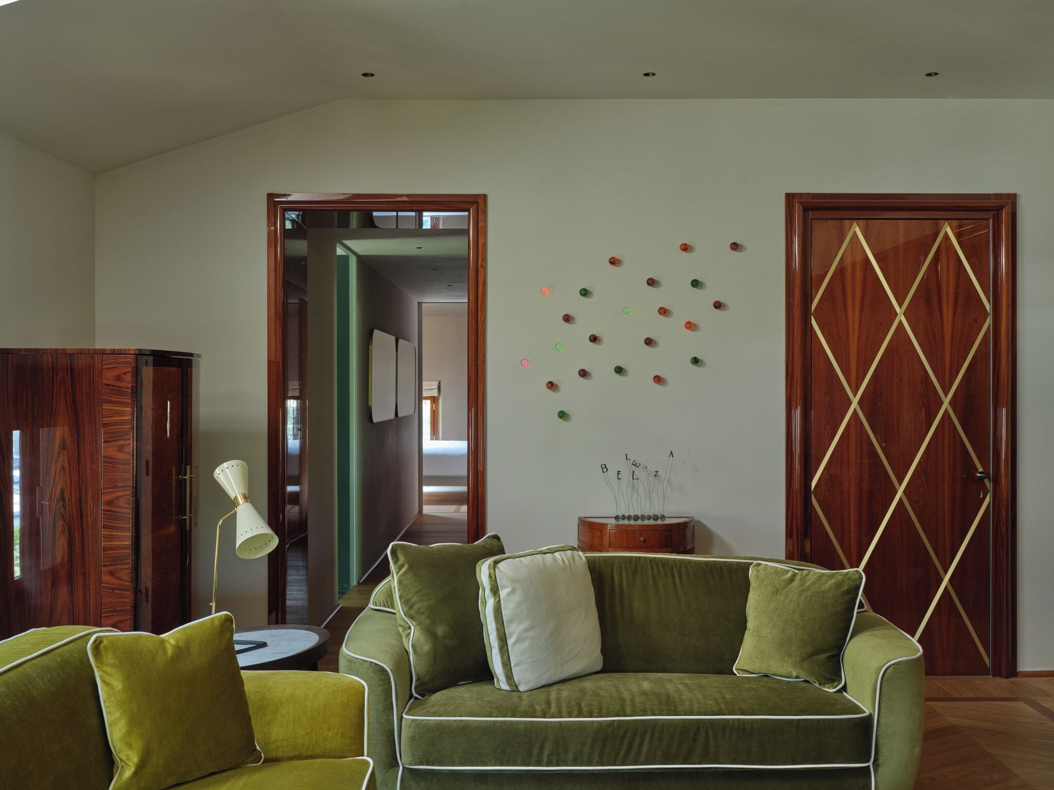 Una sala de estar con un sofá verde, una mesita redonda pequeña y una lámpara. Se observa un armario de madera y una puerta con un diseño geométrico.