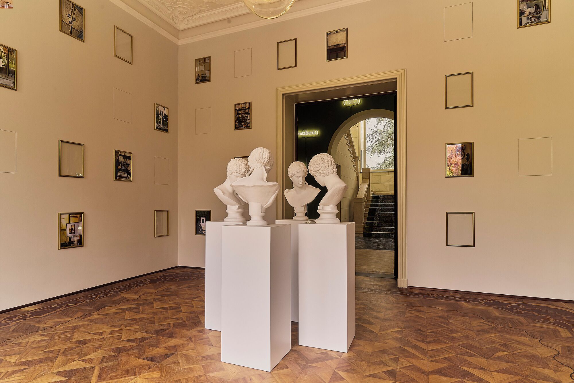 A hallway with white pedestals displaying bust sculptures, framed pictures on the walls, and a staircase leading to another area.