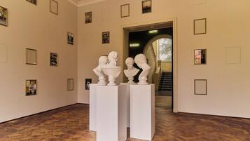 A hallway with white pedestals displaying bust sculptures, framed pictures on the walls, and a staircase leading to another area.