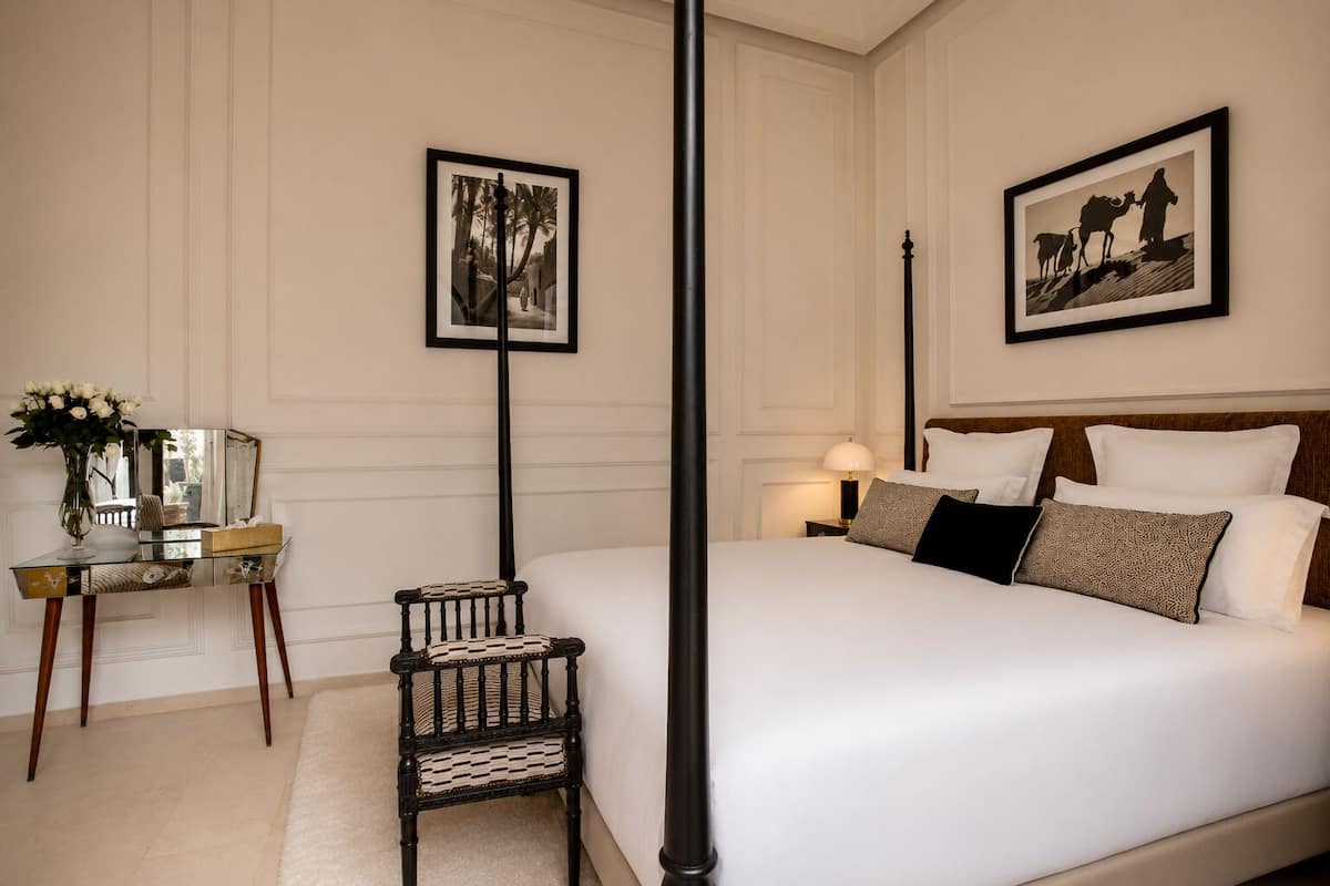 A bedroom with a four-poster bed, a nightstand with a lamp, a chair, and two framed pictures on the wall.