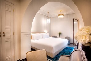 Une chambre d’hôtel avec un grand lit, un coin salon avec un fauteuil, une petite table, un miroir et un vase avec des fleurs.