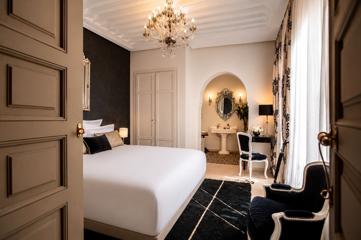 A hotel room with a large bed, a desk, a chair, a chandelier, a mirror, and a bathroom with a sink and mirror.