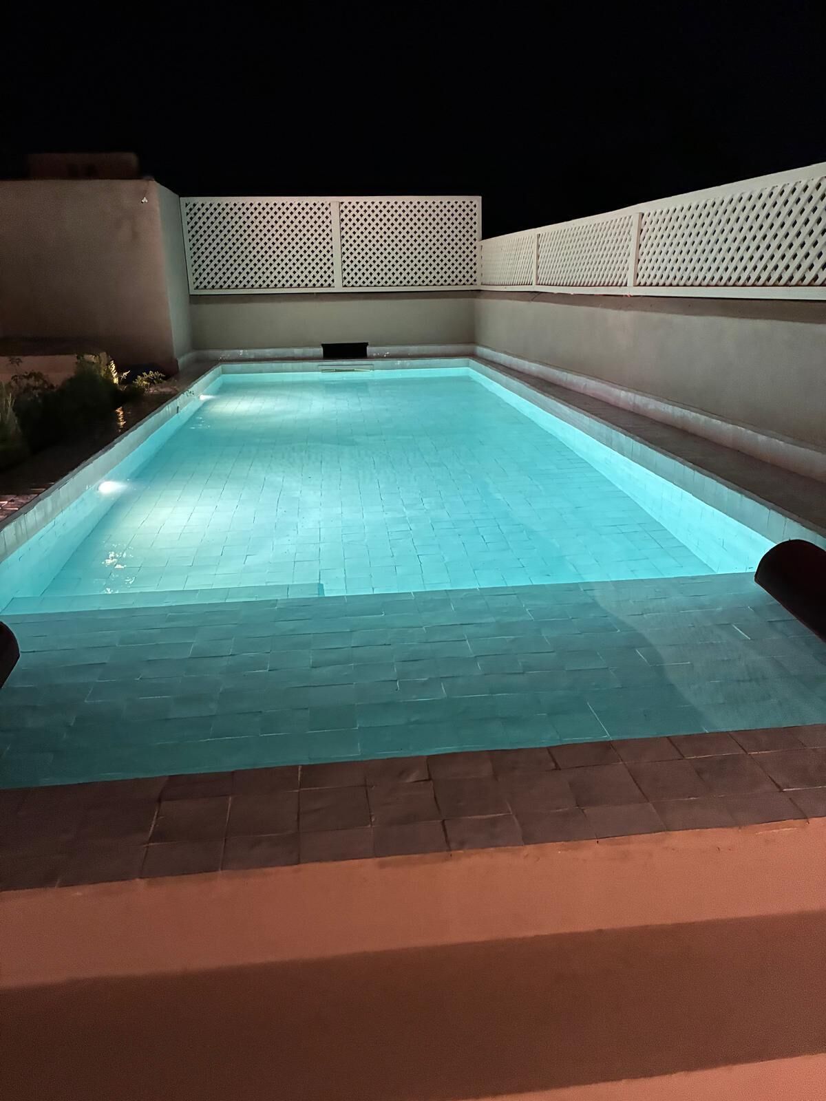 A rectangular swimming pool with a tiled deck, surrounded by walls and a fence.
