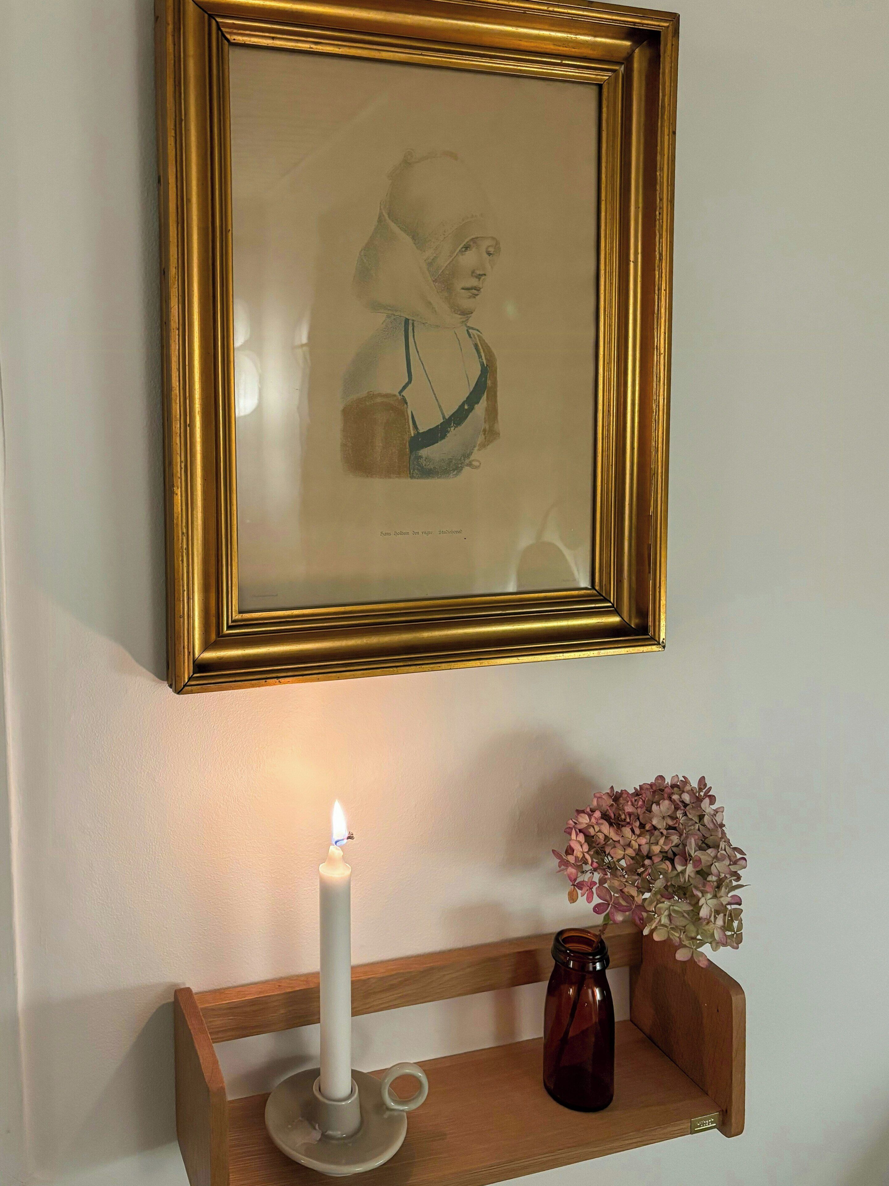 Et indrammet kunstværk på en væg, et tændt lys og en vase med blomster på en hylde.
