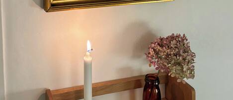 Et indrammet kunstværk på en væg, et tændt lys og en vase med blomster på en hylde.