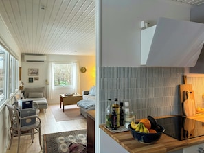 A modern living room with a sofa, a dining area with a table and chairs, and a kitchen with a cutting board and a bowl of fruit.