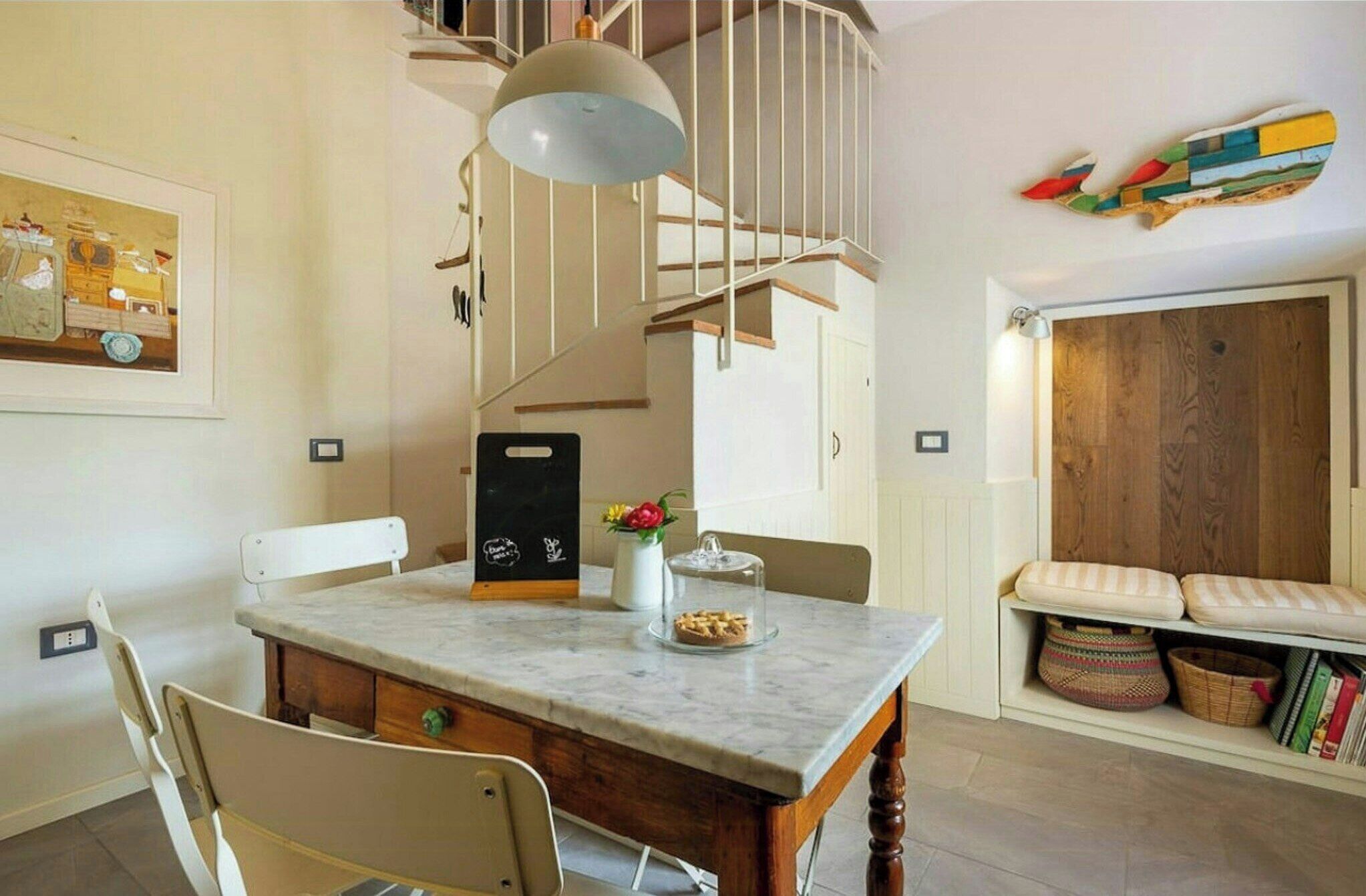 Zona de menjador amb taula de marbre, cadires, lampadari, escala i paret de fusta amb una obra d'art colorida.