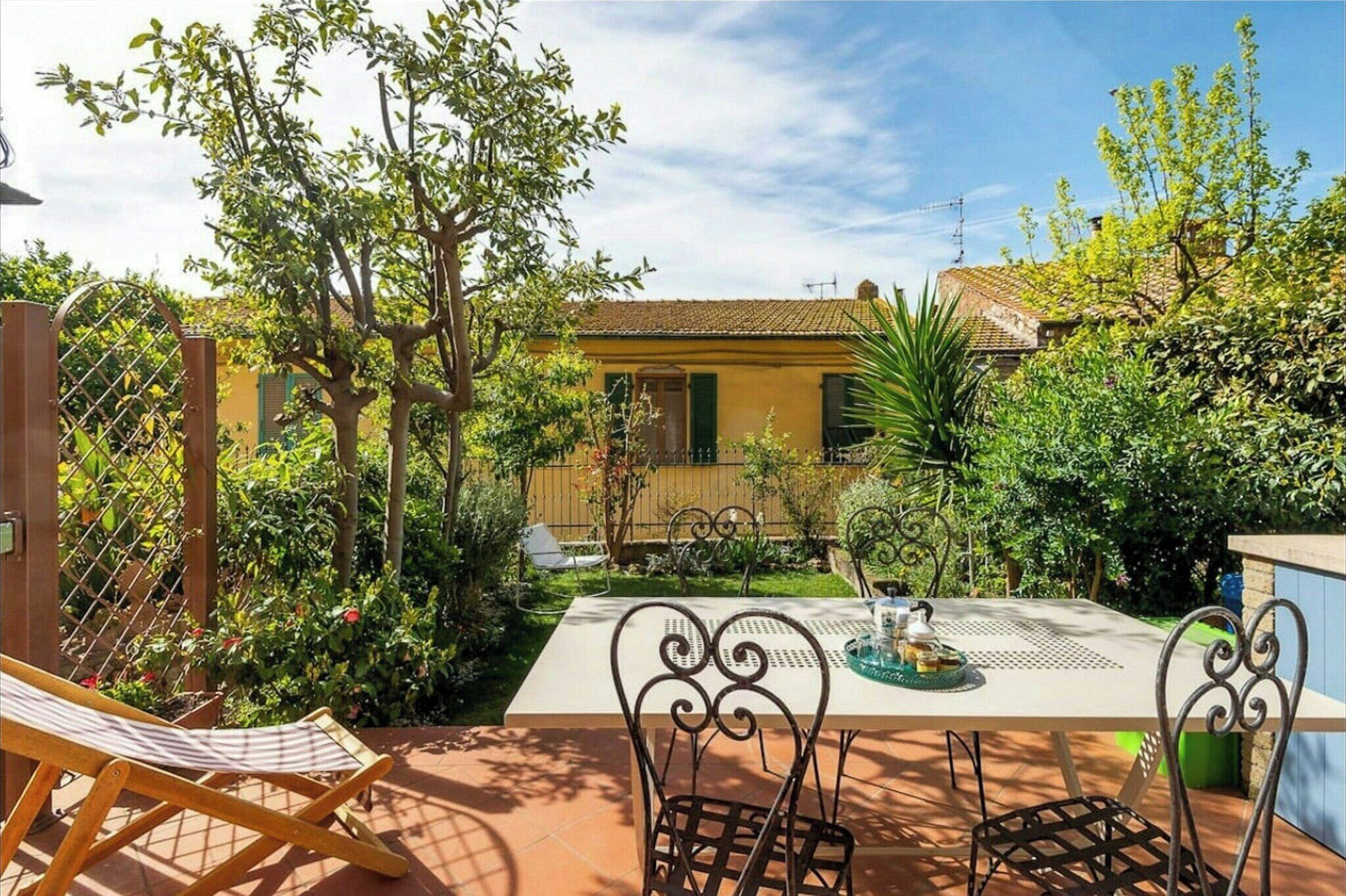 A patio with a table and chairs, surrounded by greenery and a building in the background.