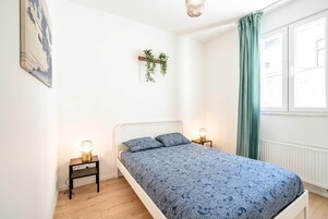 Quarto com cama, abajures, planta na parede e janela com cortinas.