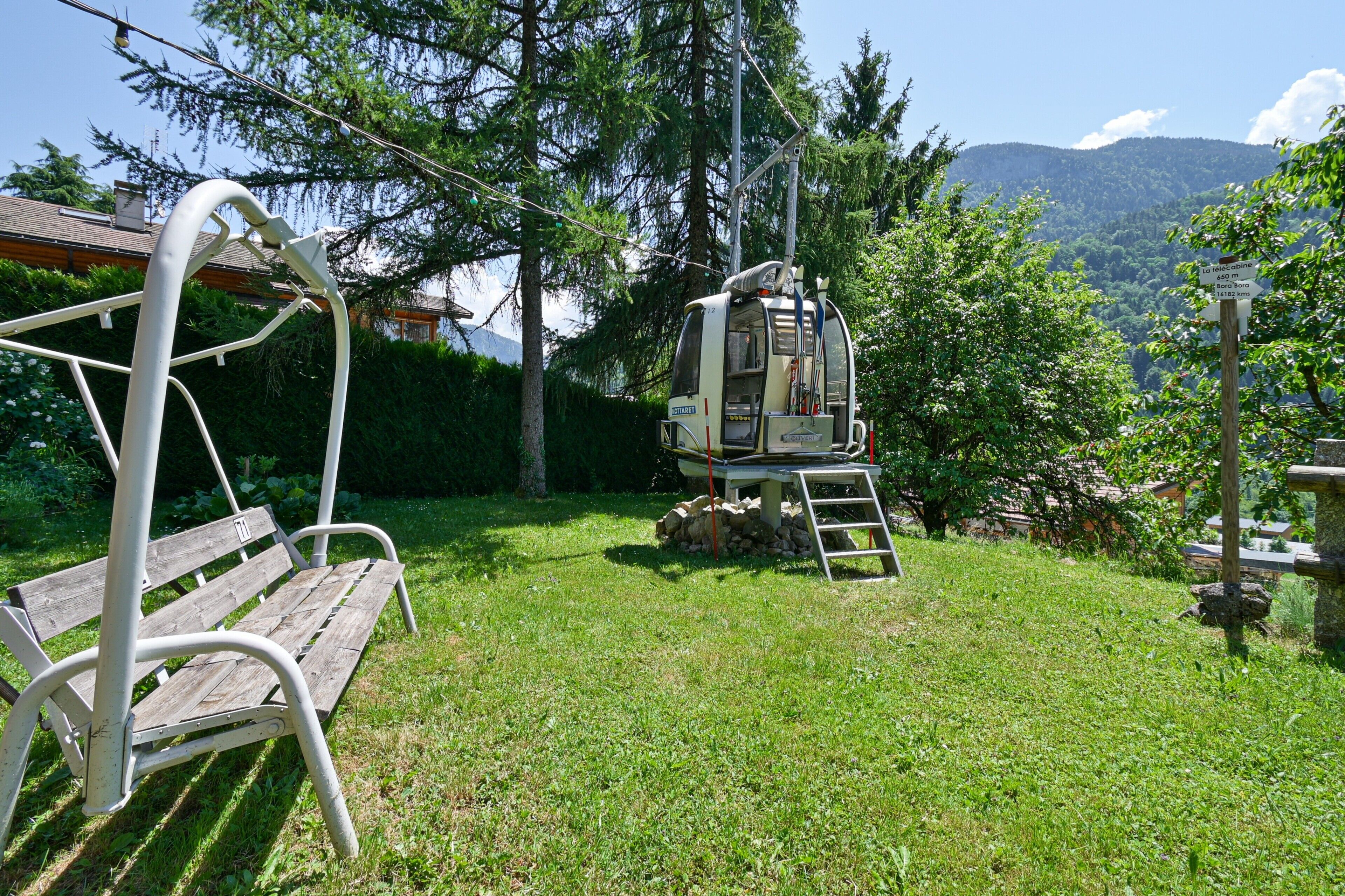 Eine hölzerne Bank, ein kleiner Tisch mit einer Stehle und eine Seilbahn in einer grünen Landschaft mit Bäumen und Bergen im Hintergrund.