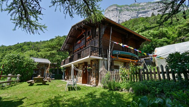 A wooden chalet with a balcony, surrounded by lush greenery and a mountain backdrop. - La Télécabine Guest Room (La Balme-de-Thuy)