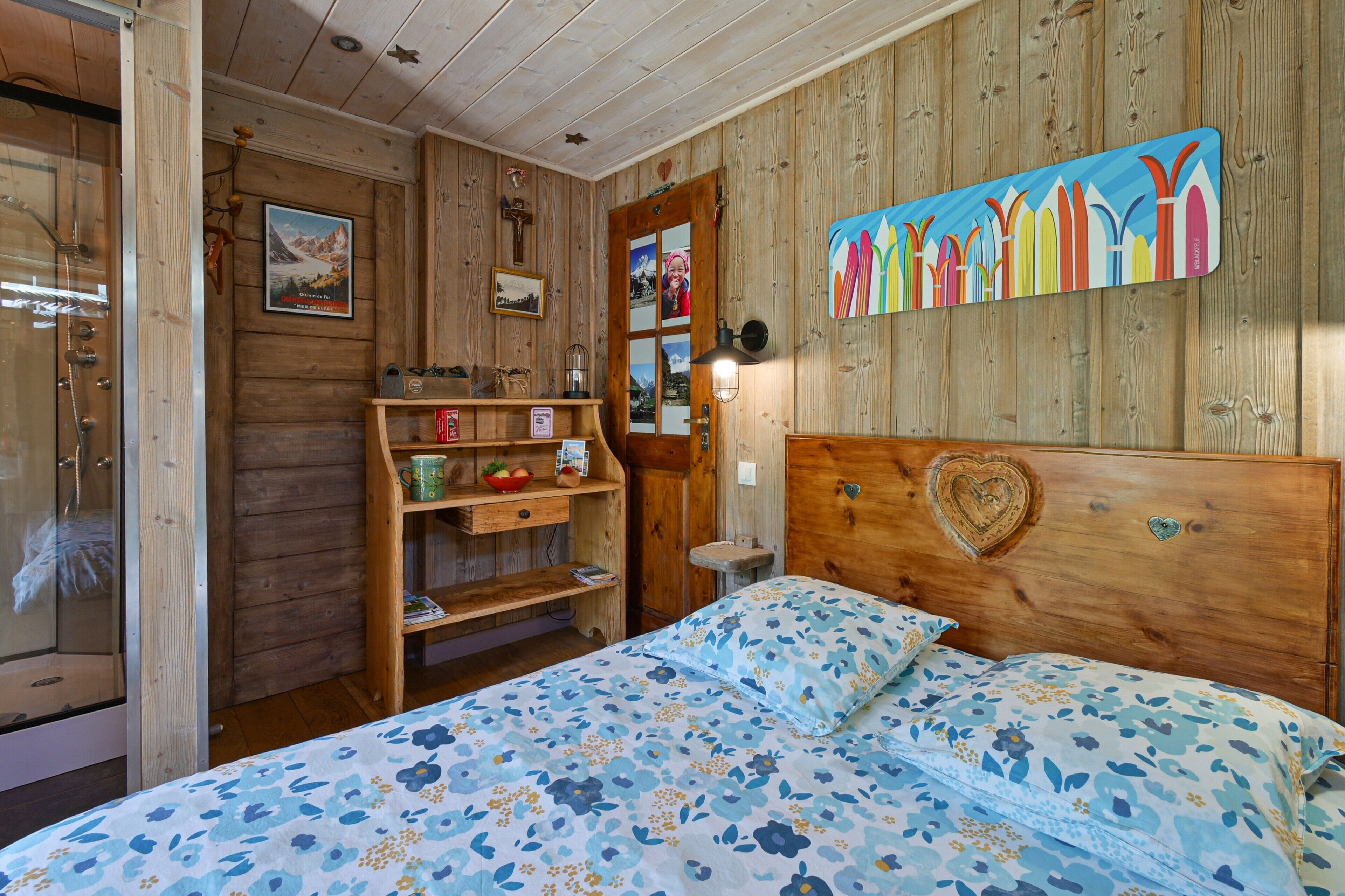 Een houten kamer met een bed, een plank met boeken en een fotolijstje, en een schilderij aan de muur.