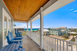 A covered balcony with wooden ceiling, blue Adirondack chairs, and a view of the beach and ocean.