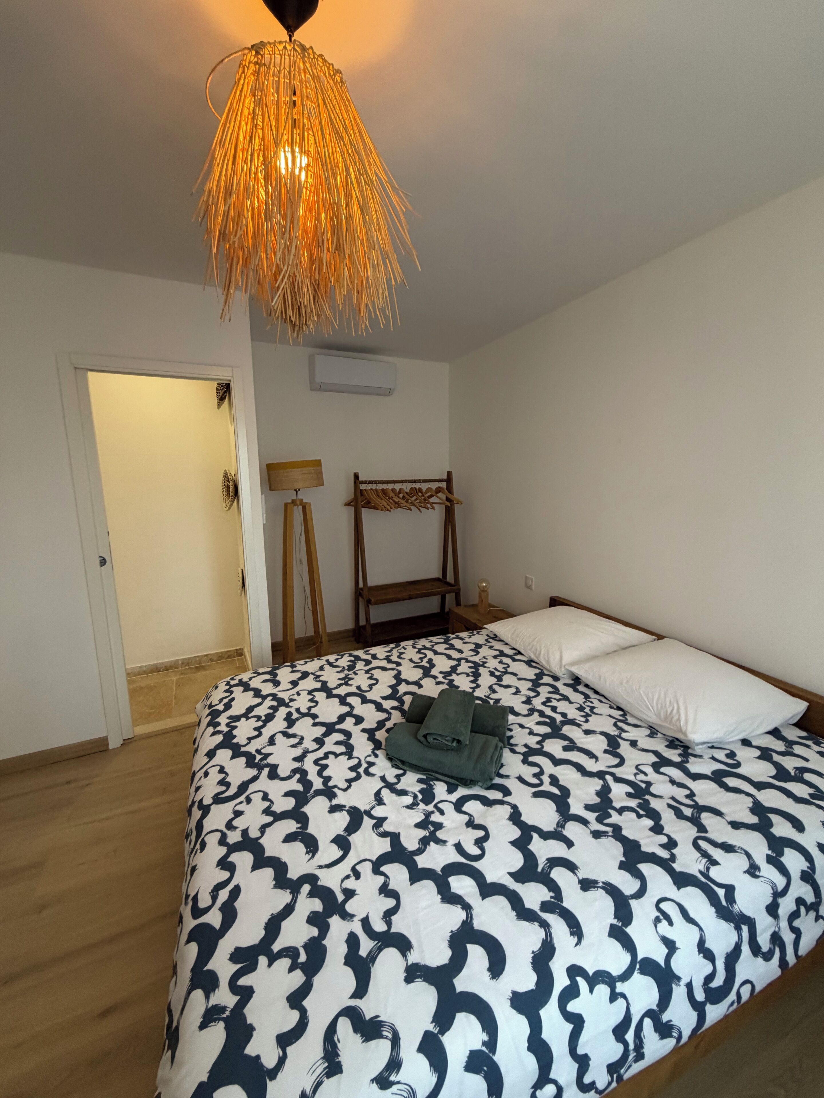 Un dormitorio con edredón estampado, lámpara colgante, perchero de madera y puerta que da a otra habitación.