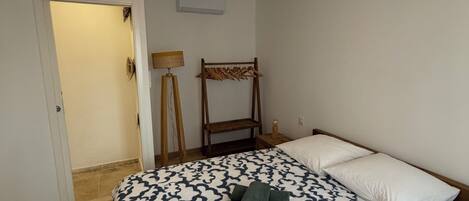 Un dormitorio con edredón estampado, lámpara colgante, perchero de madera y puerta que da a otra habitación.