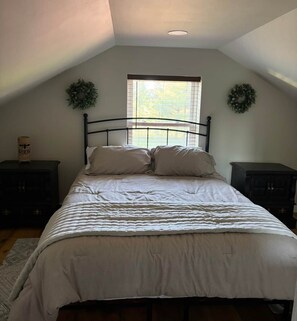 Un dormitorio con una cama, dos mesitas de noche, un ventanal con persianas y dos coronas decorativas en la pared.