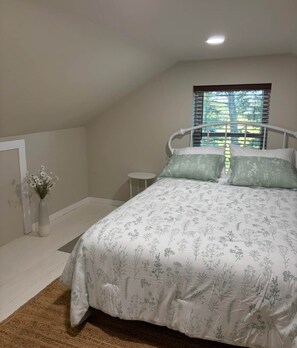 A bedroom with a bed, a small bedside table, a vase with flowers, and a window with blinds.