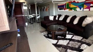 Uma sala de estar moderna com um sofá marrom escuro, uma mesa de centro de vidro e um quadro abstrato colorido na parede.